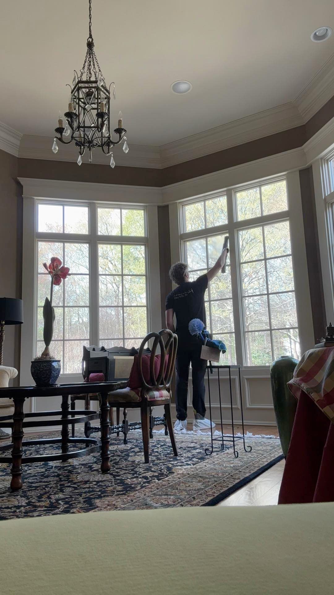 A person on a ladder cleaning a window in a bright room with a chandelier.