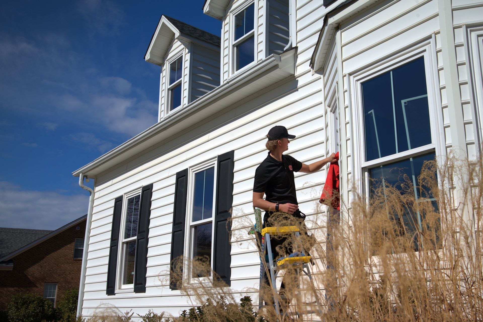 Person on a ladder cleaning a window on a white house with black shutters, on a sunny day.