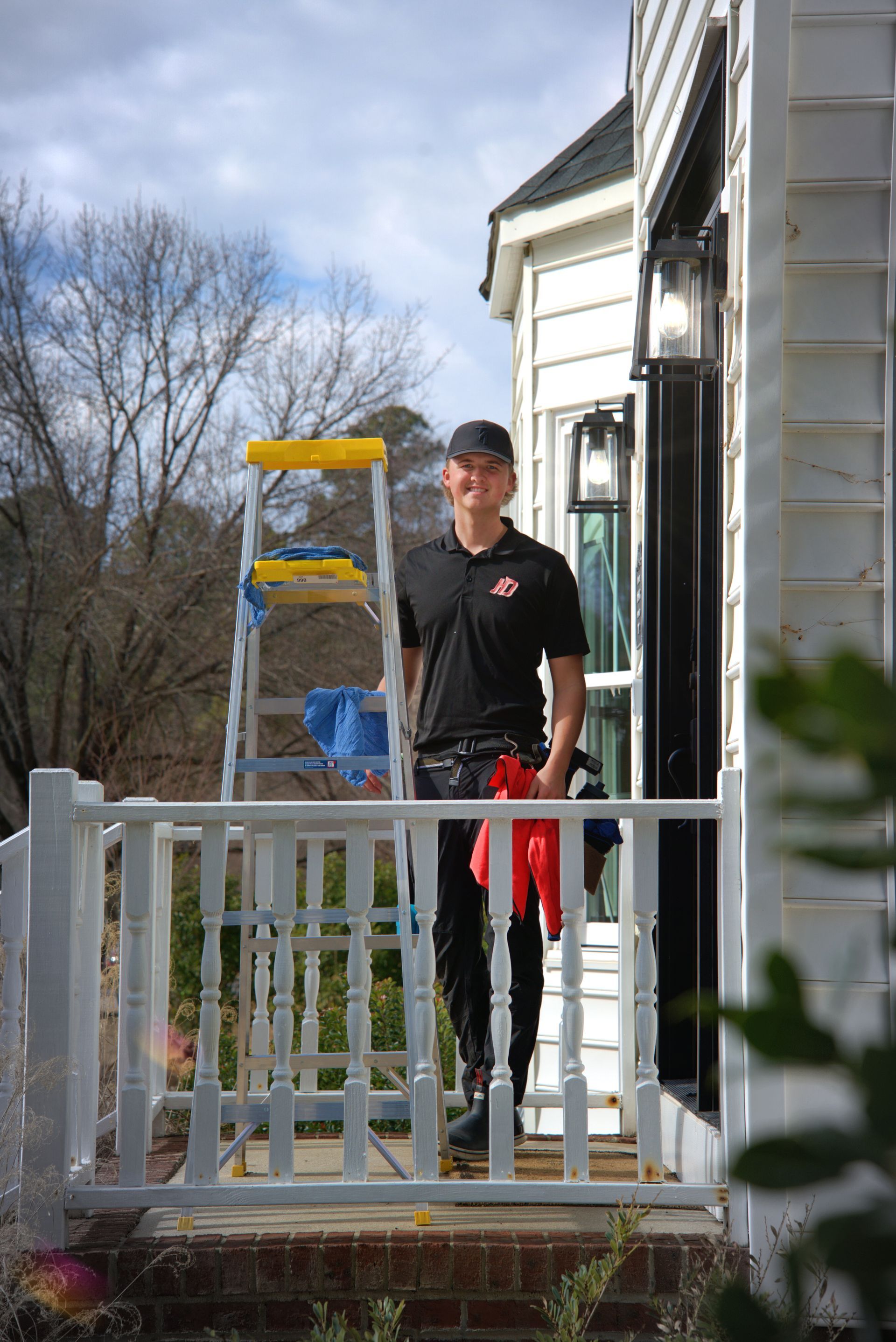 Window cleaner on a porch, smiling. Ladder with cleaning supplies nearby. Building with white siding in background.