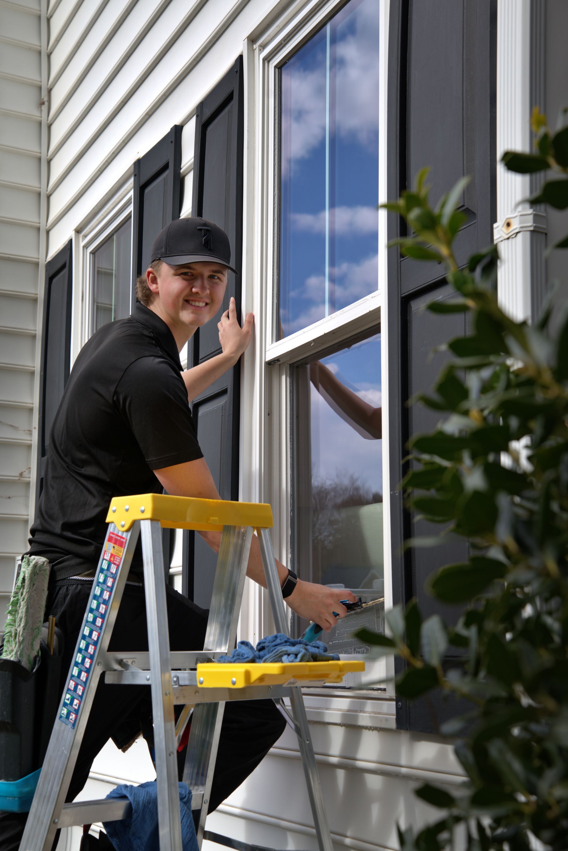 Man cleaning a window on a house exterior, smiling. Standing on a stepladder, using a squeegee.