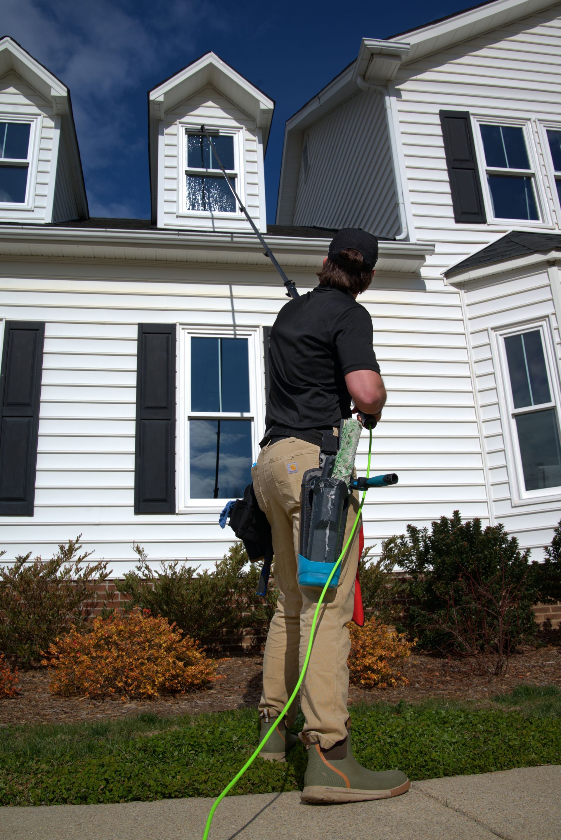 Window cleaner using a pole to wash windows on a white house, standing on a sidewalk.