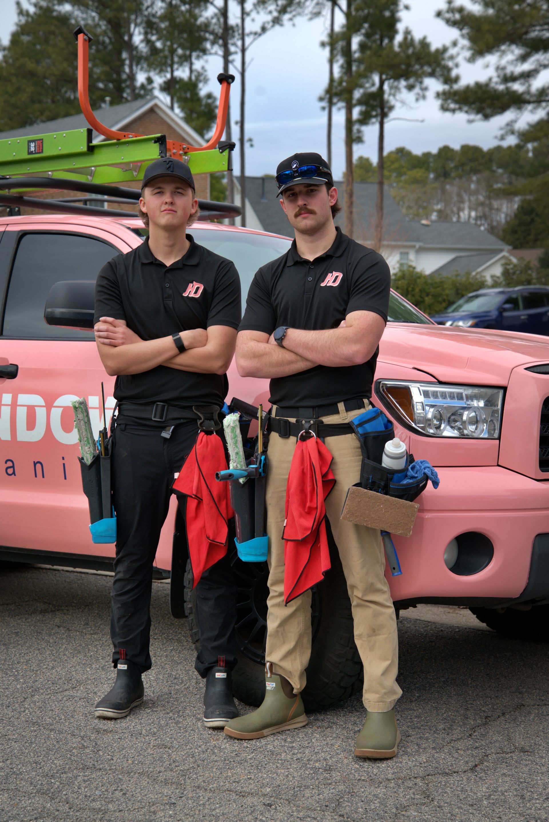 Two window cleaners stand in front of a pink truck, arms crossed. They wear black shirts, hats, and tool belts.