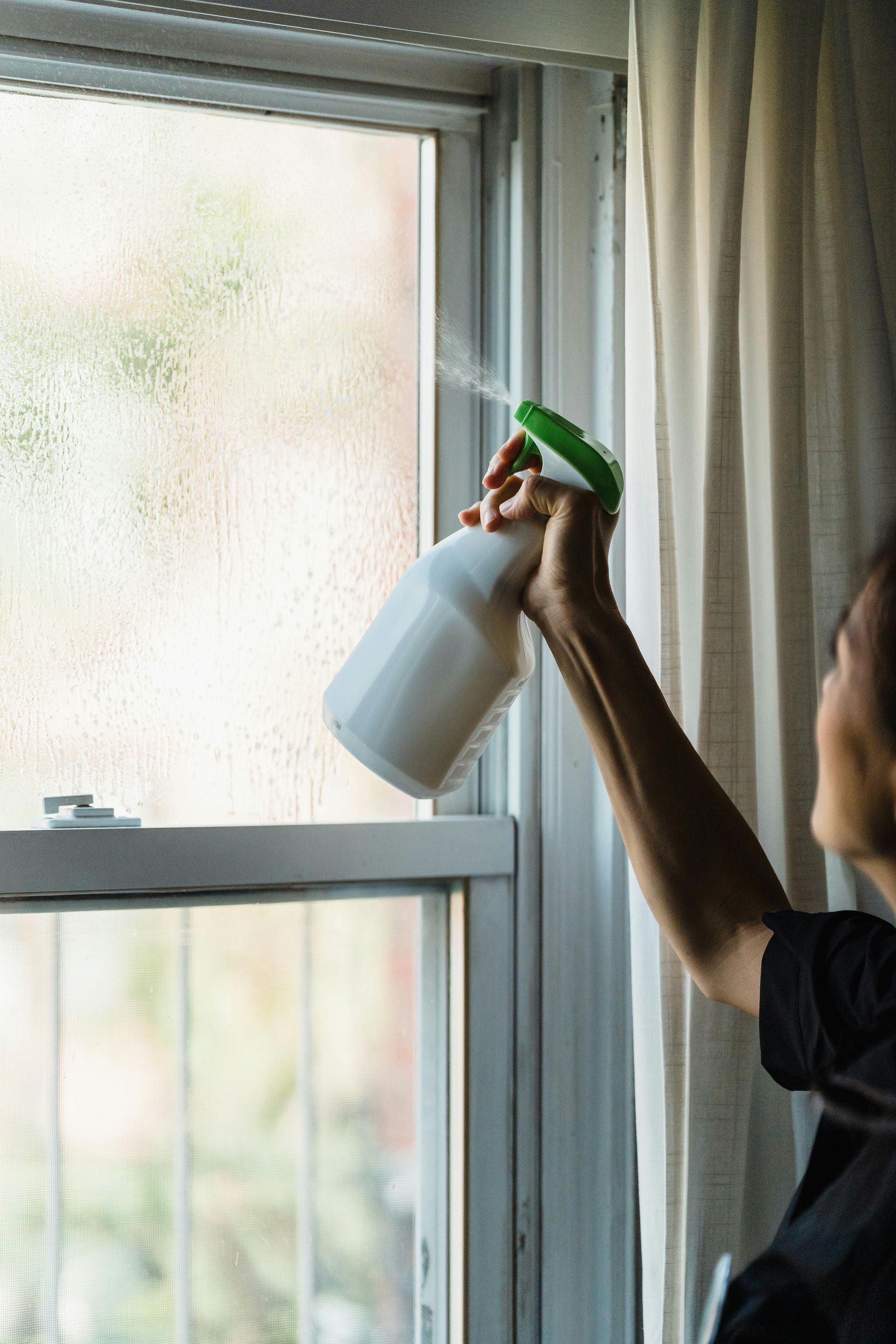 Person spraying a window with a white spray bottle; window is slightly fogged.