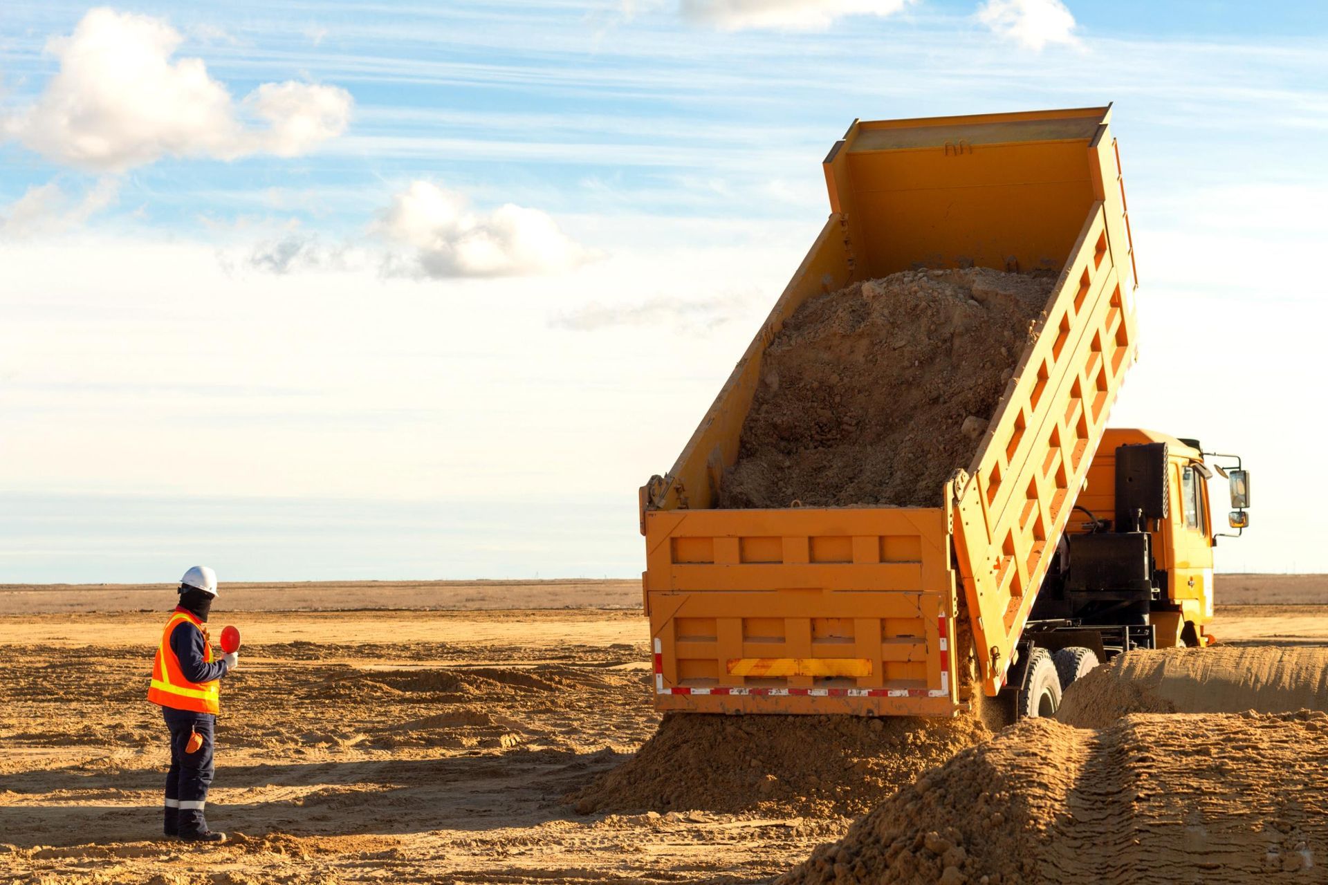 A man is standing next to a dump truck filled with dirt.