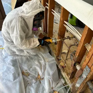 Worker in white protective suit using a drill to remove damaged wall studs in a stripped interior room.