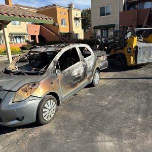 Burned silver sedan being towed by a skid steer on a residential street near apartment buildings