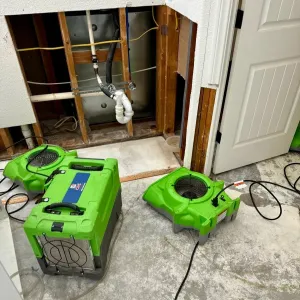 Two bright green dehumidifiers on a basement floor beside an open wall with exposed plumbing.