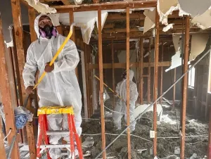 Two workers in protective suits demolish interior walls in a damaged room.