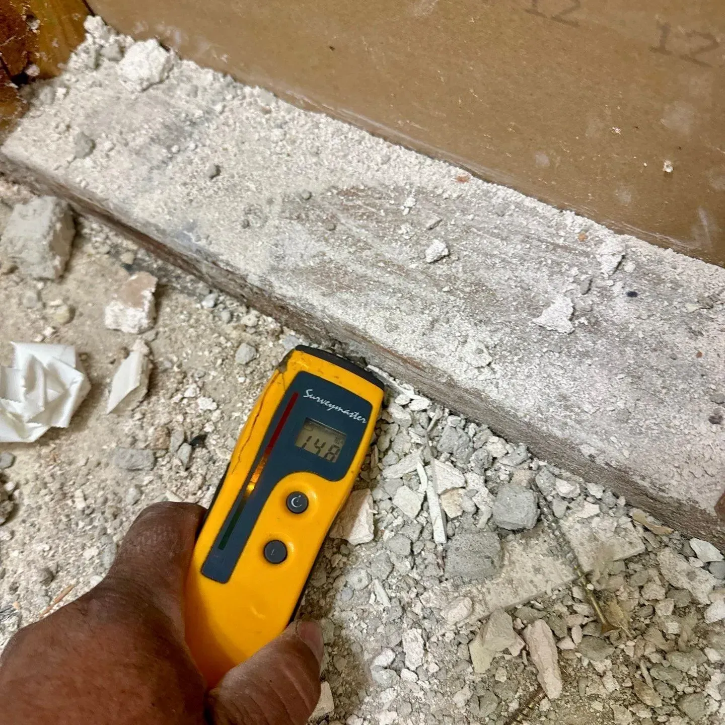 Hand holding a yellow infrared thermometer over dusty debris beside a wall