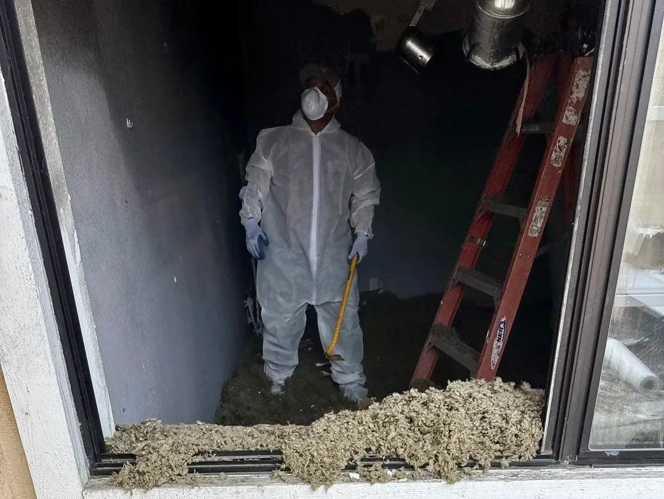 Person in protective suit standing in a dark doorway beside a ladder and debris spill