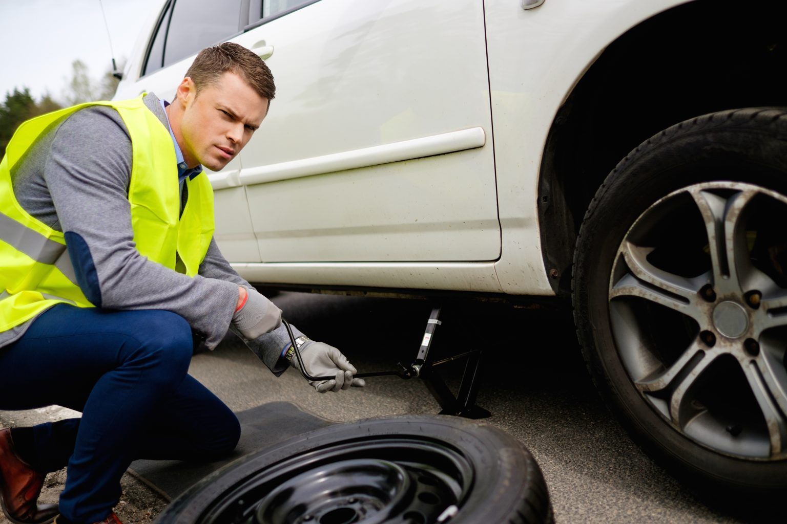 Man in safety vest changing a tire on a white car, outdoors.
