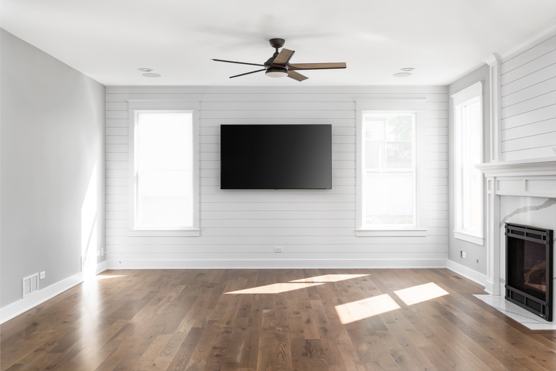 An empty living room with hardwood floors , a fireplace , a flat screen tv and a ceiling fan.