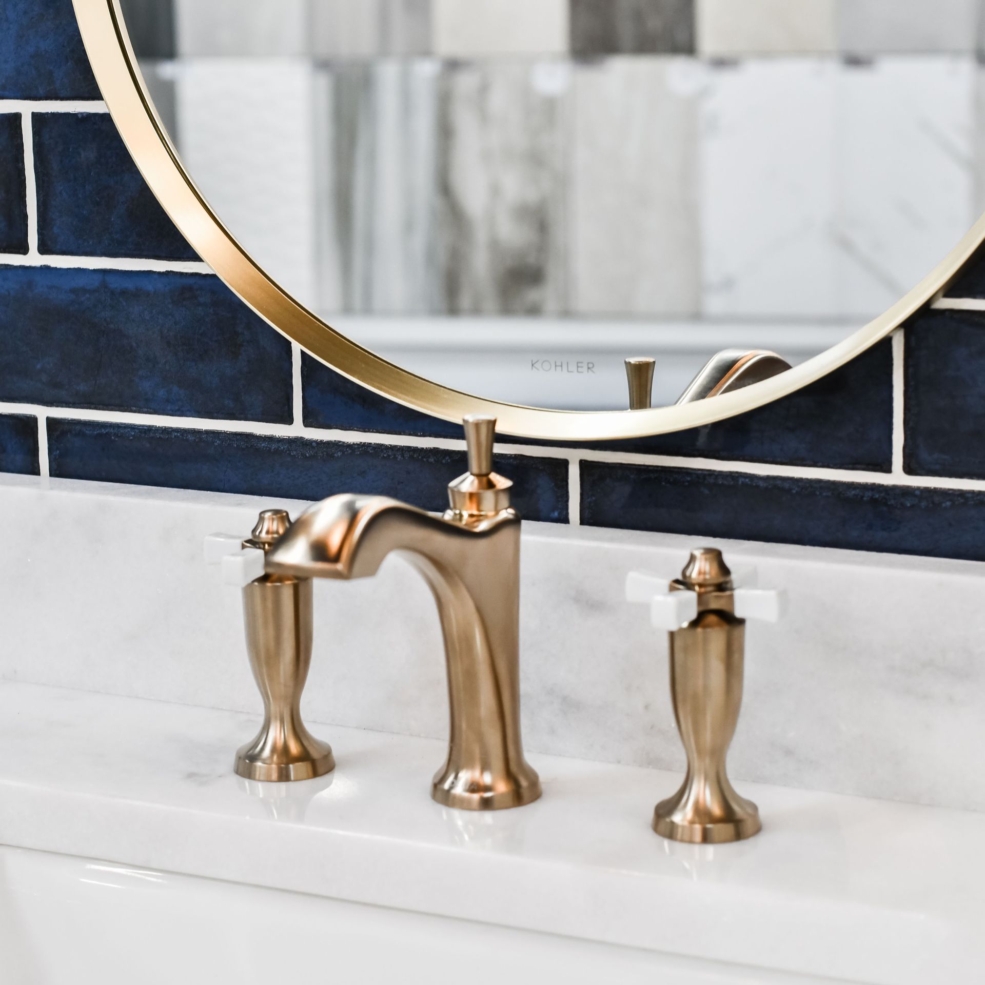 A modern brass bathroom faucet with white cross handles sits on a white marble vanity against deep blue subway tiles.
