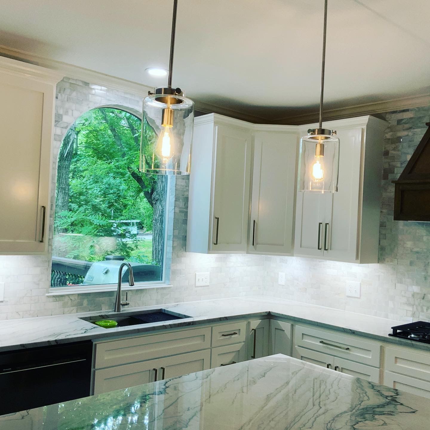 A modern kitchen featuring white cabinets, a stone island countertop, two glass pendant lights, and an arched window view.