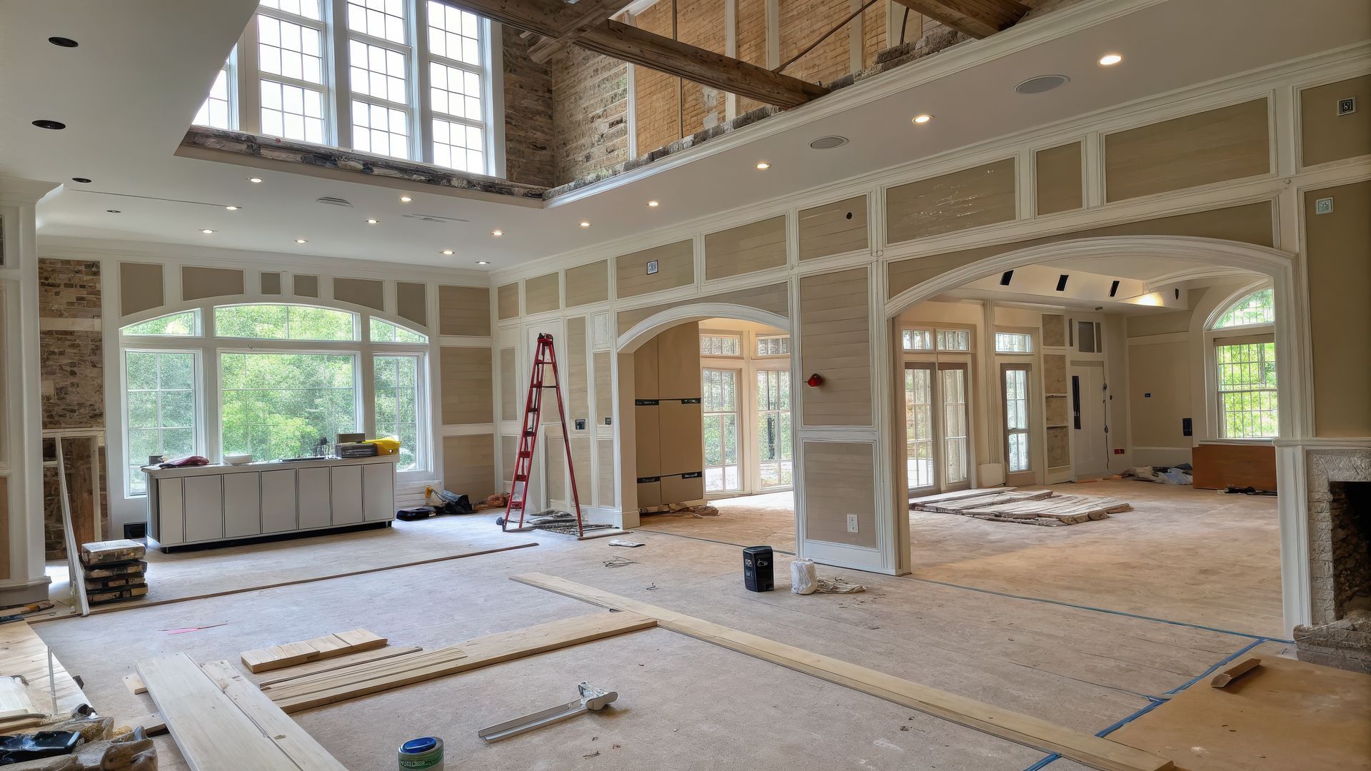 A large room under construction with white trim, unfinished walls, arched doorways, large windows, and a red step ladder.