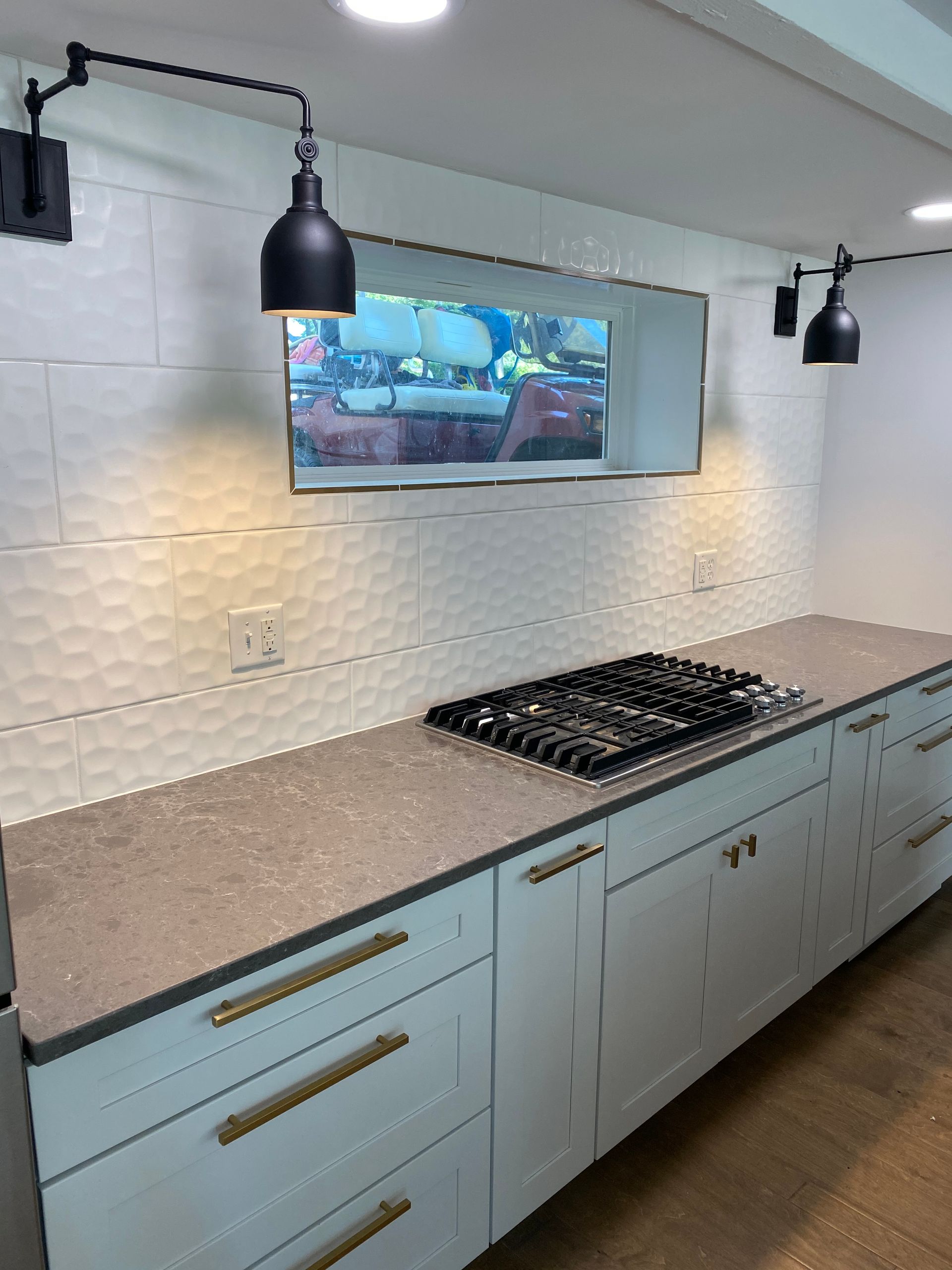 Modern kitchen featuring light blue cabinets with brass pulls, a grey quartz countertop, gas cooktop, and two black sconces.