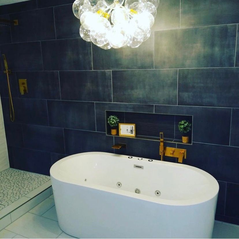 A modern bathroom featuring a white soaking tub against a dark tiled wall with a recessed shelf, gold fixtures, and light.