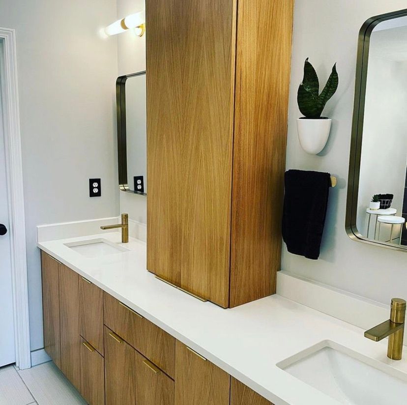 A modern bathroom vanity with wood cabinets, white countertops, dual gold faucets, two mirrors, and a wall-mounted plant.