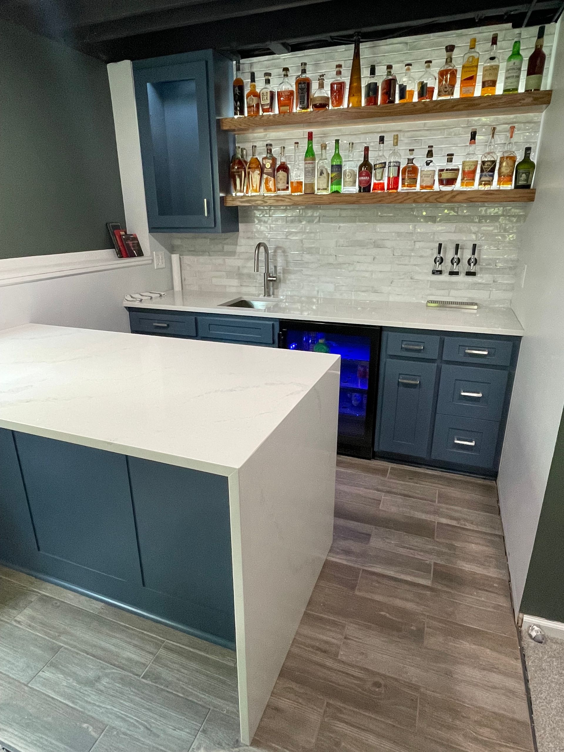 A modern home bar with blue cabinets, a quartz island, wood shelves holding liquor bottles, and a tile backsplash.