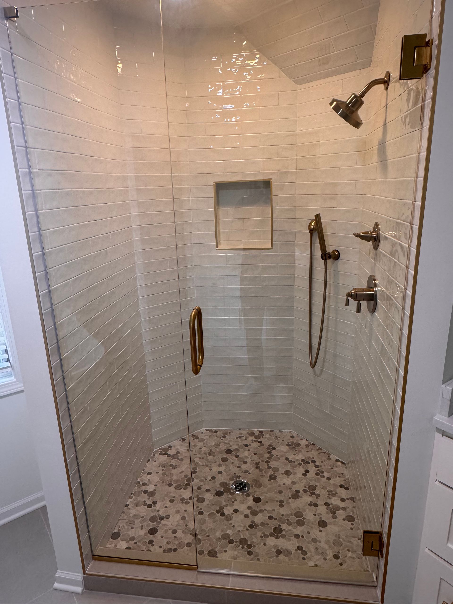 A glass shower enclosure with white tiled walls, gold fixtures, a hand-held shower head, and a stone pebble floor.