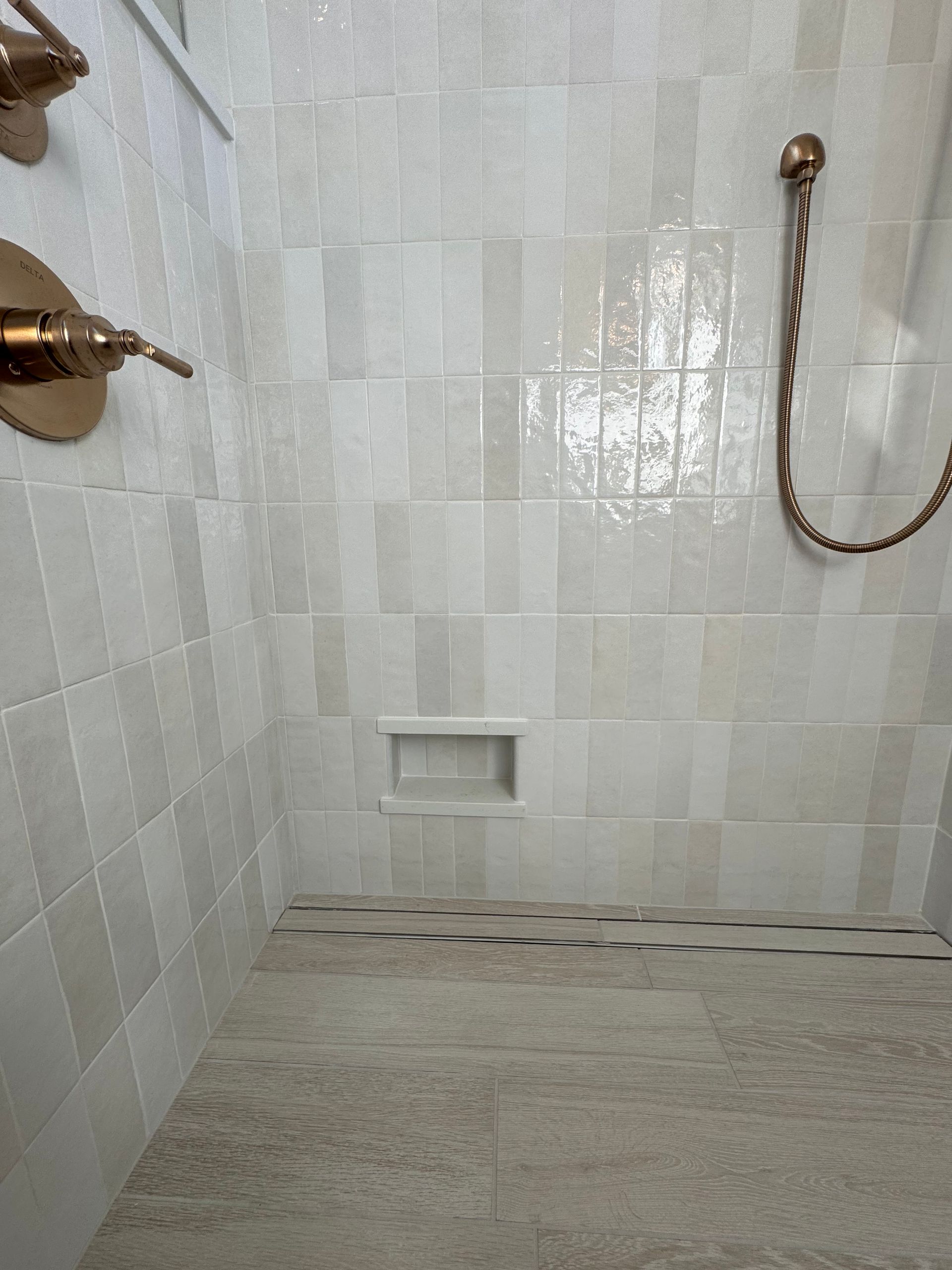 A shower interior with vertical, light-colored glossy tiles, a small recessed niche, and rose gold fixtures.