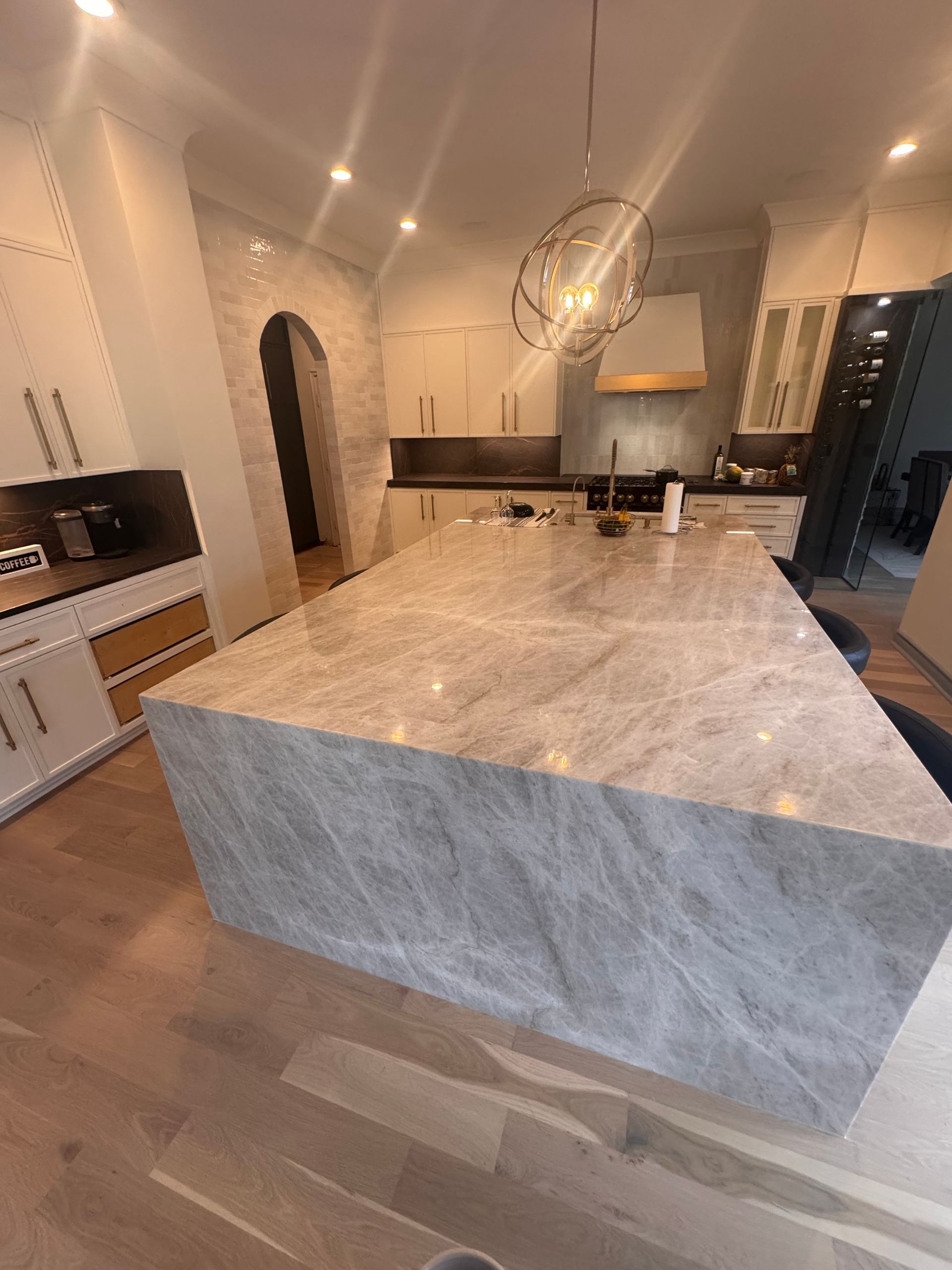 A large, light-colored stone kitchen island with a waterfall edge sits in a modern kitchen with wood floors.