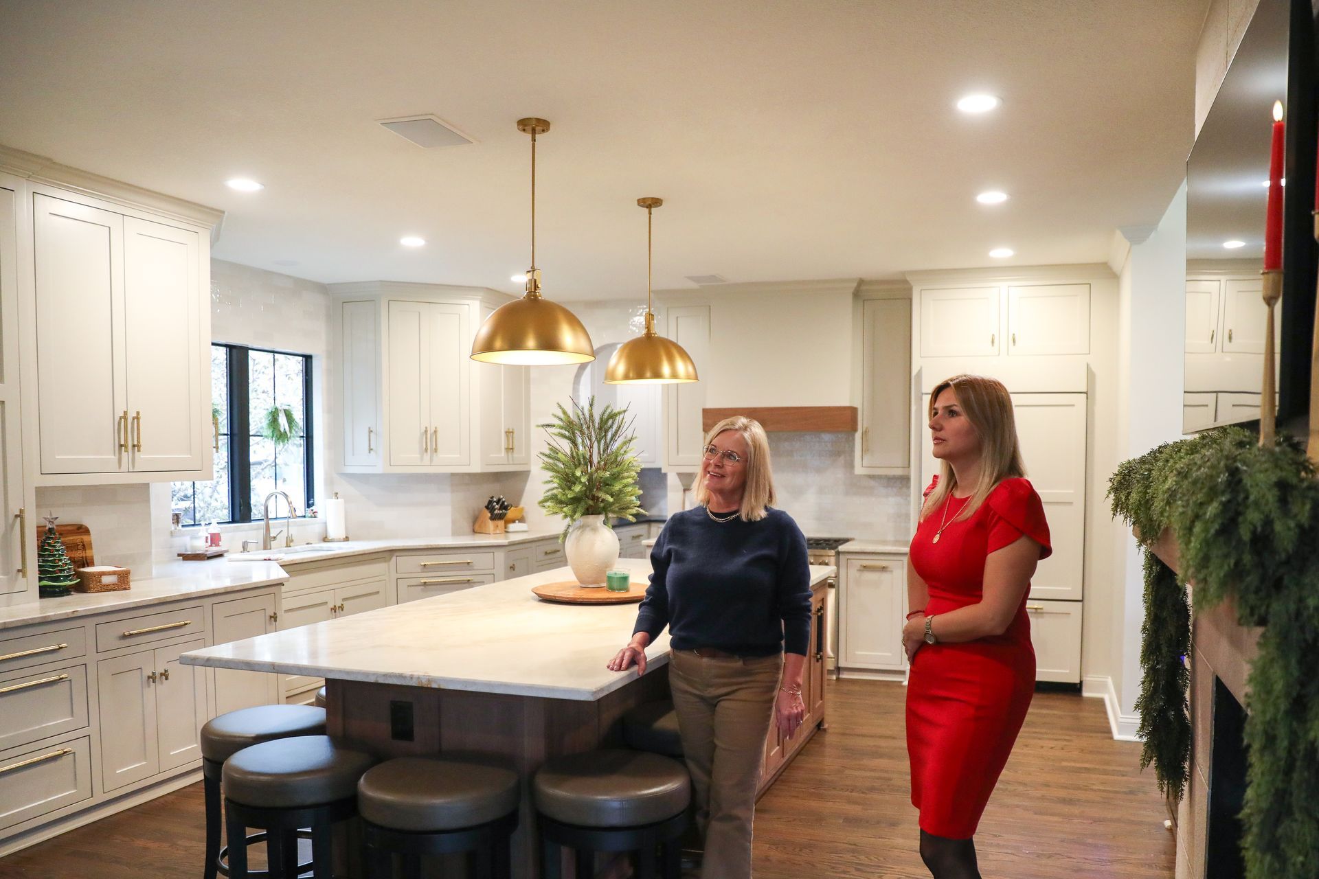 Two people stand in a spacious kitchen with white cabinets, a large central island, and two brass pendant lights.