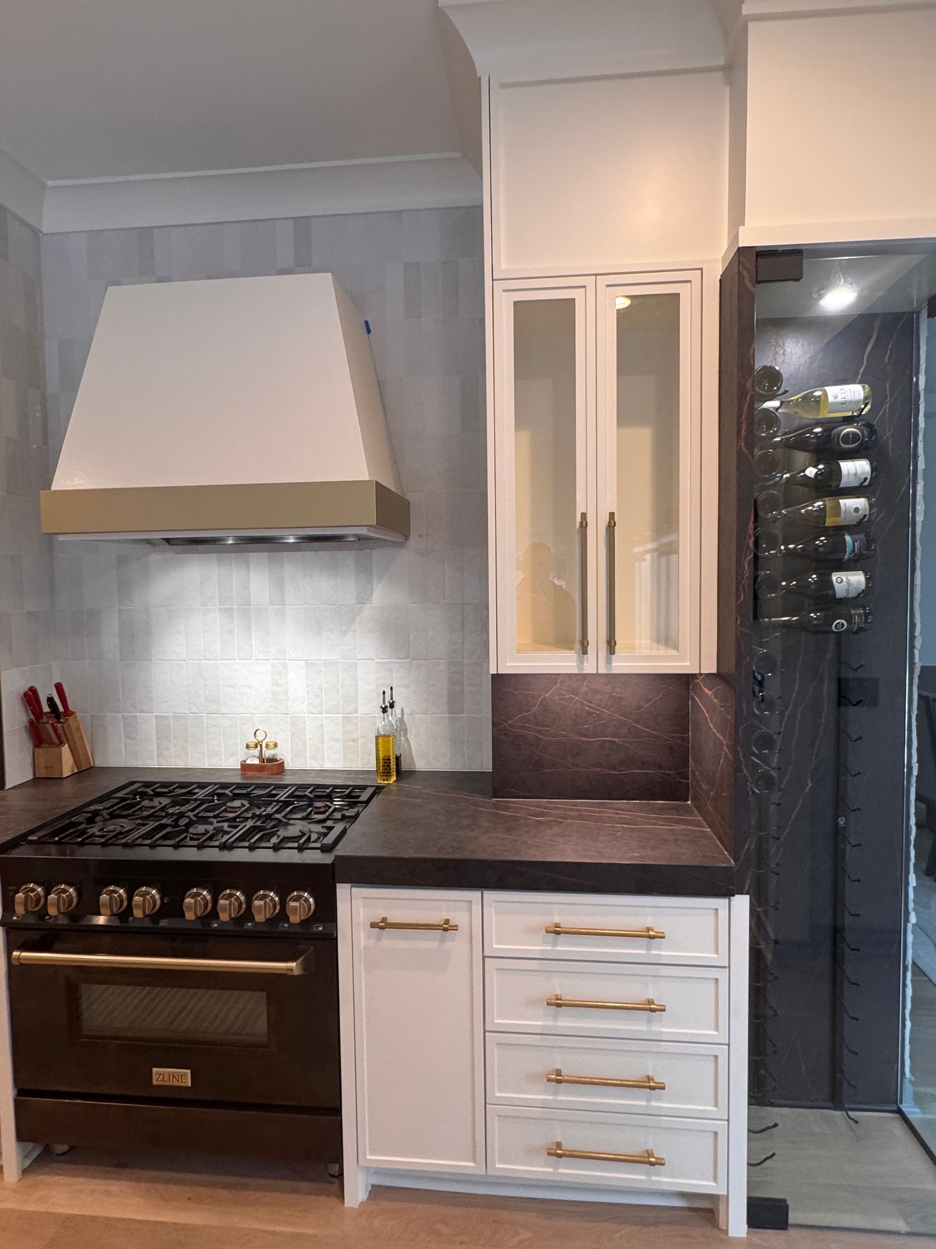 A kitchen featuring a black stove, white cabinets with gold hardware, a white range hood, and a vertical wine rack.