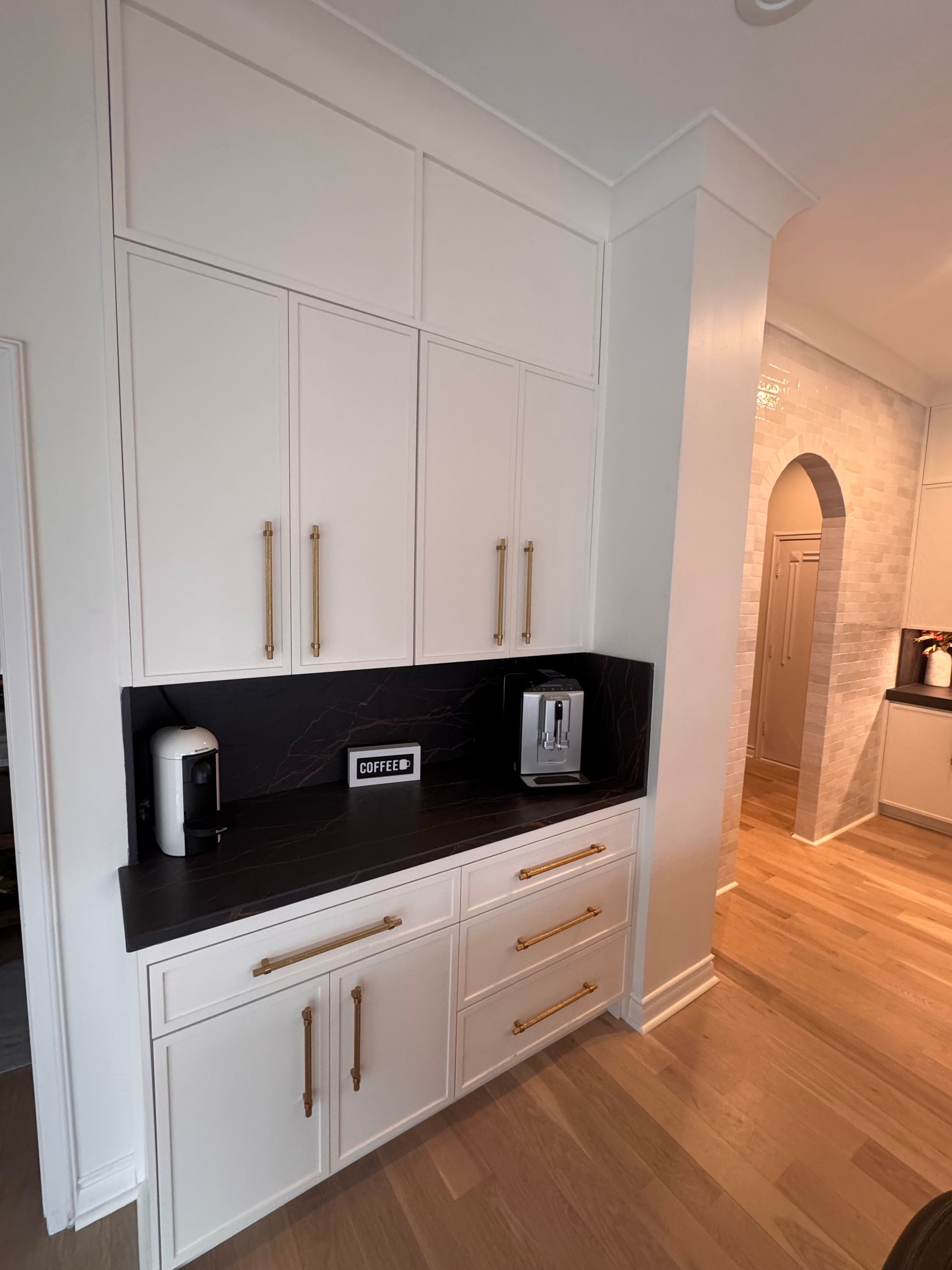 White cabinetry with brass hardware and a black countertop, featuring coffee appliances in a home kitchen setting.