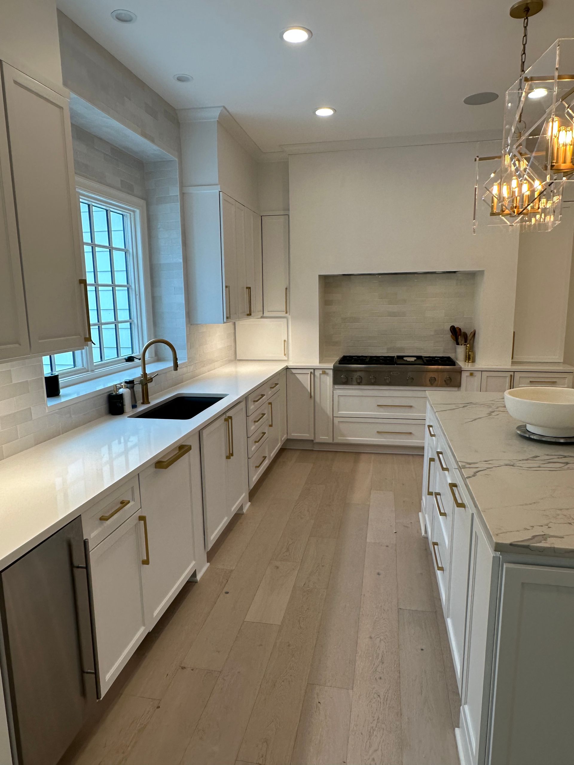 A bright, modern kitchen featuring white shaker cabinets, marble countertops, light wood floors, and a hanging chandelier.