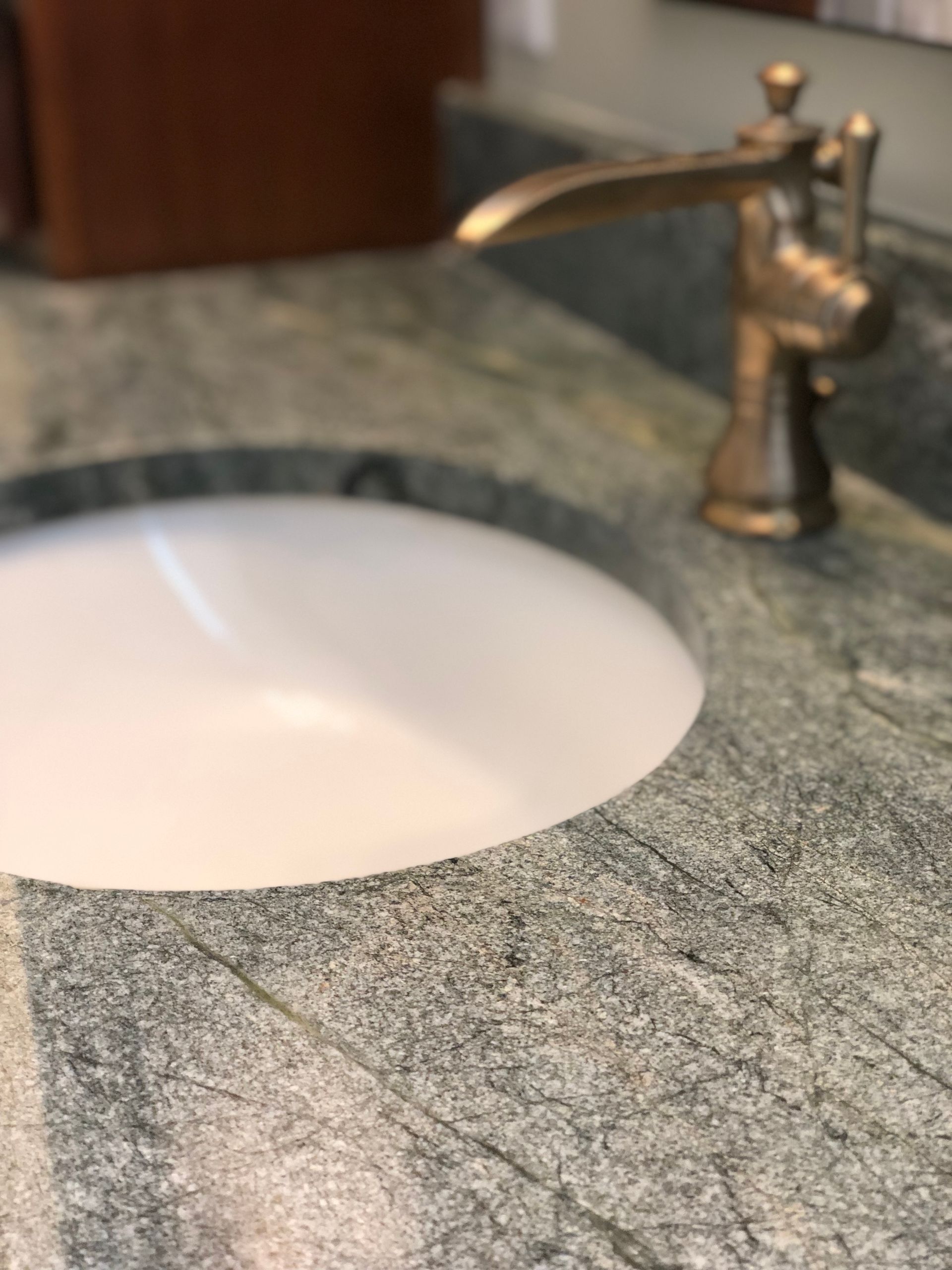 A white undermount bathroom sink set in a grey granite countertop with a bronze-toned, single-handle faucet.