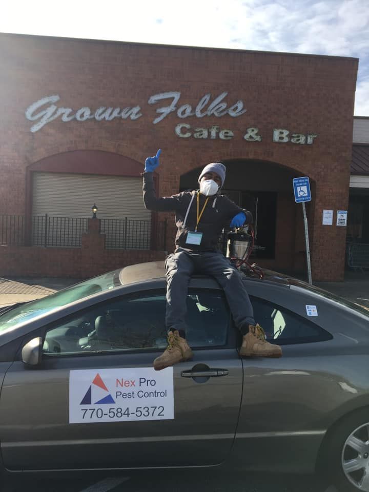A man wearing a mask is sitting on the hood of a car in front of a grown folks cafe and bar.