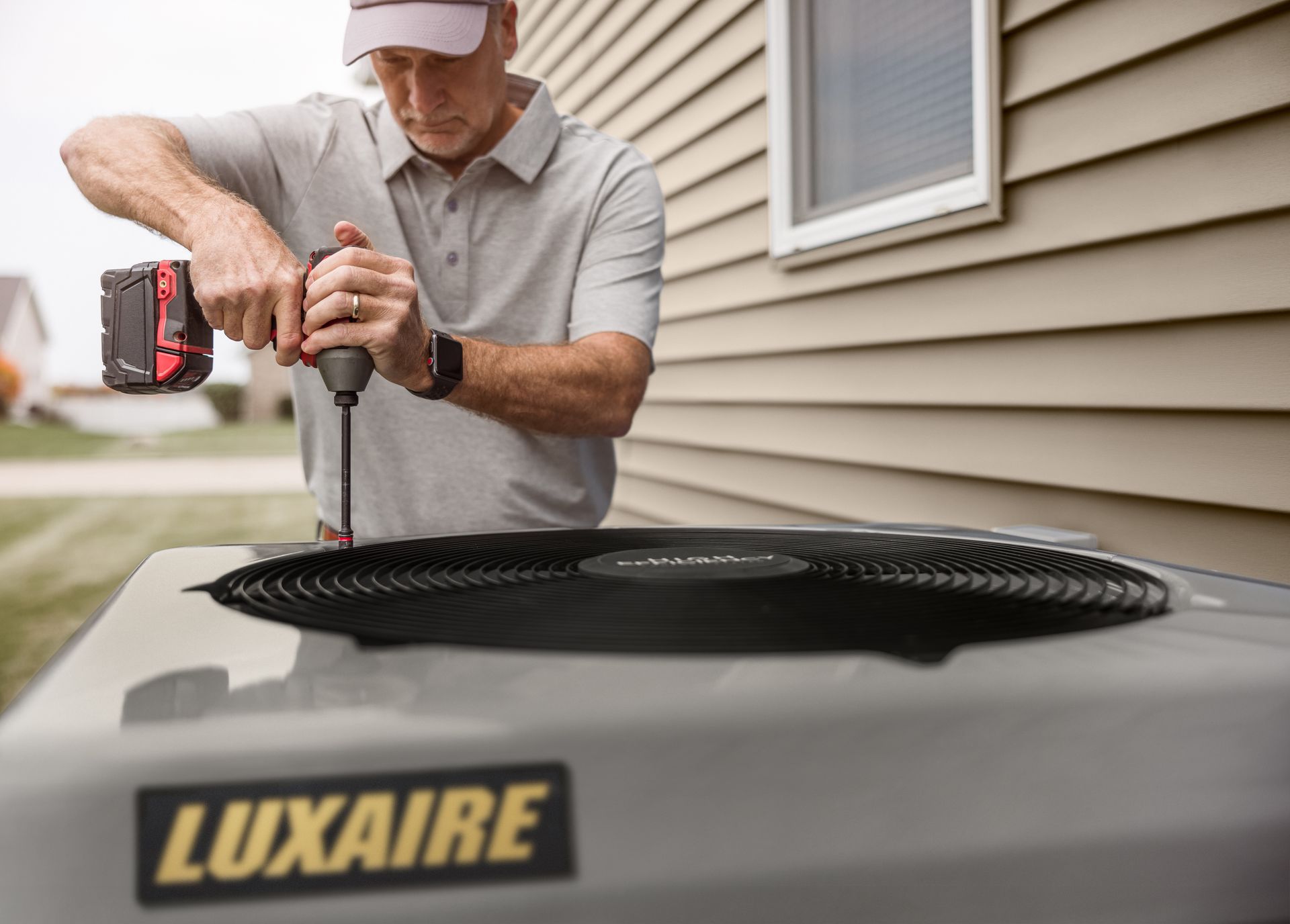 A gray outdoor air conditioning condenser unit sits on a concrete pad against a brick wall, with a small bush nearby.