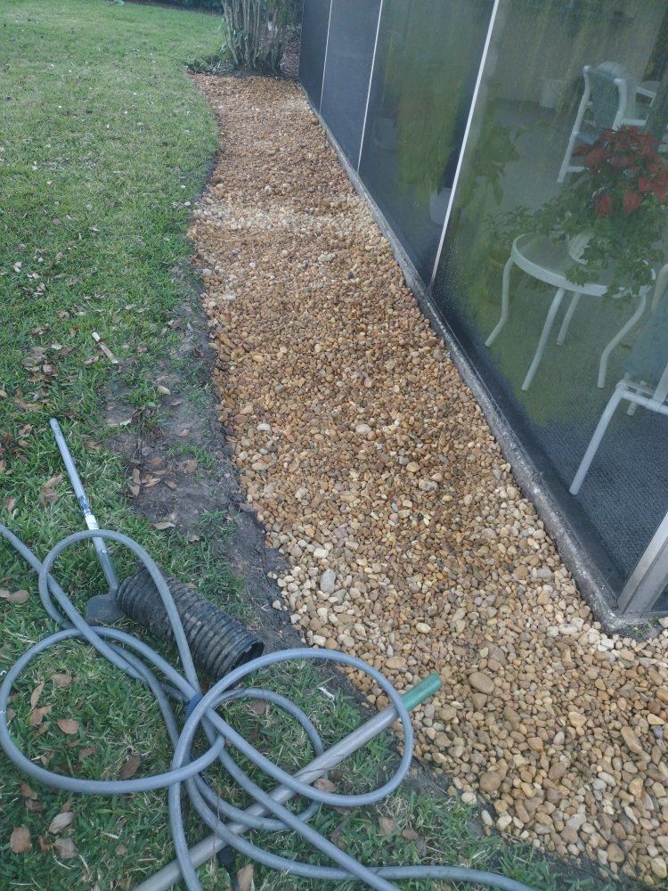 A gravel path lined with tan rocks runs along the base of a screened-in porch next to a grassy lawn and a garden hose.