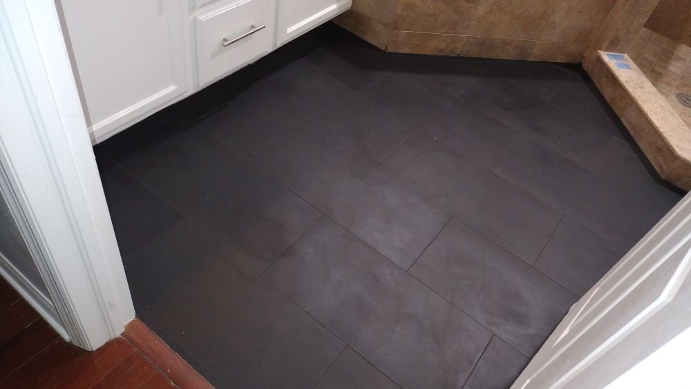 A bathroom floor with dark, rectangular slate-style tiles extending from a white cabinet to a tiled shower ledge.