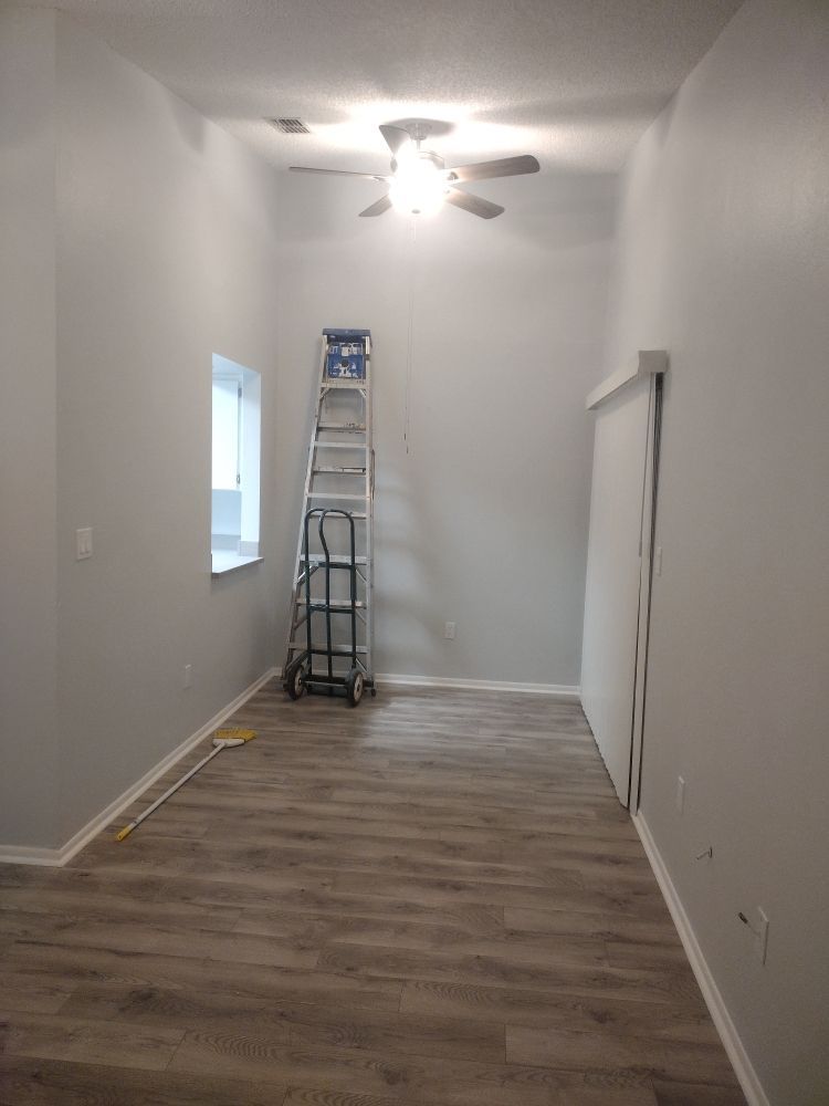 An empty, grey-walled room with wood-look flooring, a ceiling fan, a window, a folding ladder, and vertical blinds.