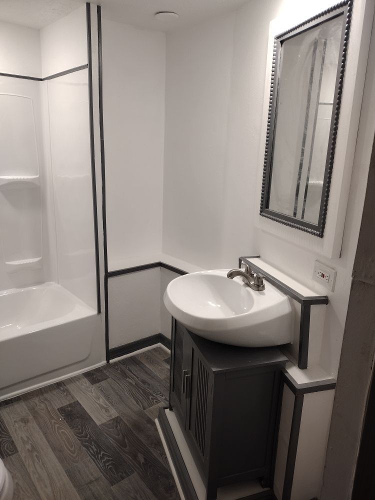 A modern bathroom featuring a white pedestal sink, a framed mirror, a bathtub, and dark grey wood-look flooring.