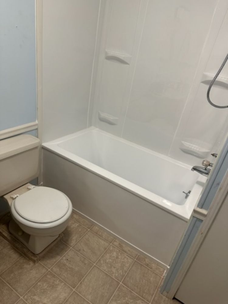 A white toilet next to a white bathtub with a shower head in a bathroom with tan tiled floors and light blue walls.