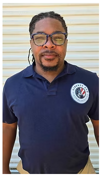 Man wearing glasses and a blue polo shirt with a company logo, in front of a neutral background.