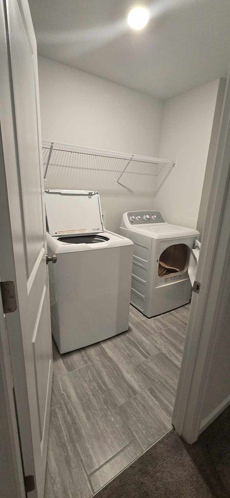 A laundry room with a washing machine, dryer, and drying rack. Gray wood-look flooring. White walls.