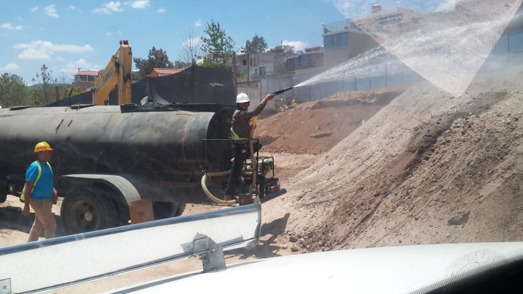 Trabajadores rocían agua sobre una gran pila de arena desde un camión cisterna de agua en un sitio de construcción.