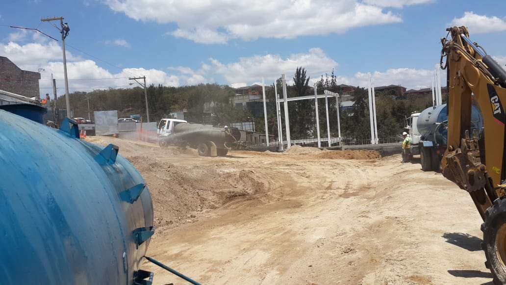 Una obra en construcción con una excavadora, camiones y un gran tanque azul. Camino de tierra polvoriento y un día soleado.
