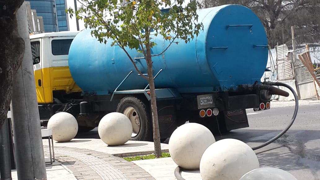 Camión de agua con tanque azul estacionado en la calle junto a barreras de bolas de concreto blancas.