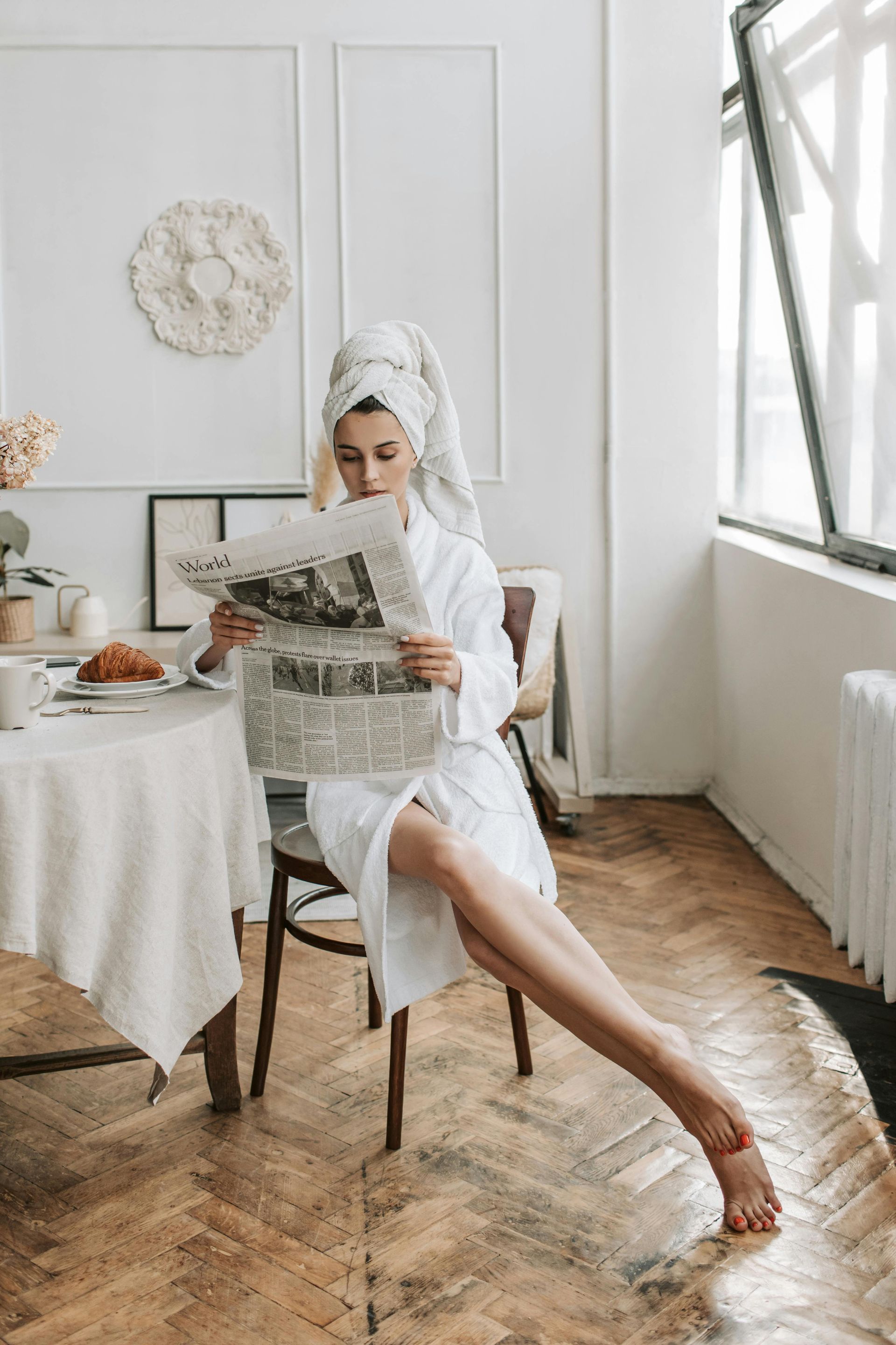 A woman in a bathrobe is sitting at a table reading a newspaper.