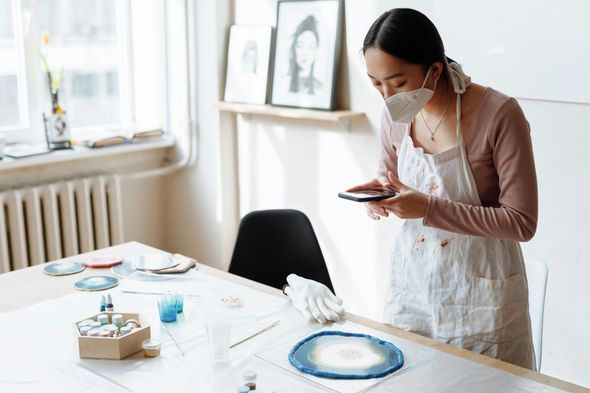 A woman wearing a mask is standing at a table looking at her phone.