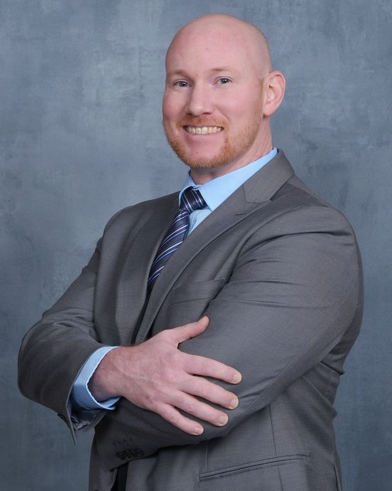 A smiling person with a shaved head wears a gray suit, light blue shirt, and patterned tie, posing with arms crossed.