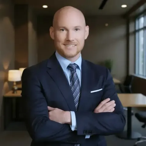 A bald professional in a navy suit and patterned tie poses with arms crossed in a modern office space.