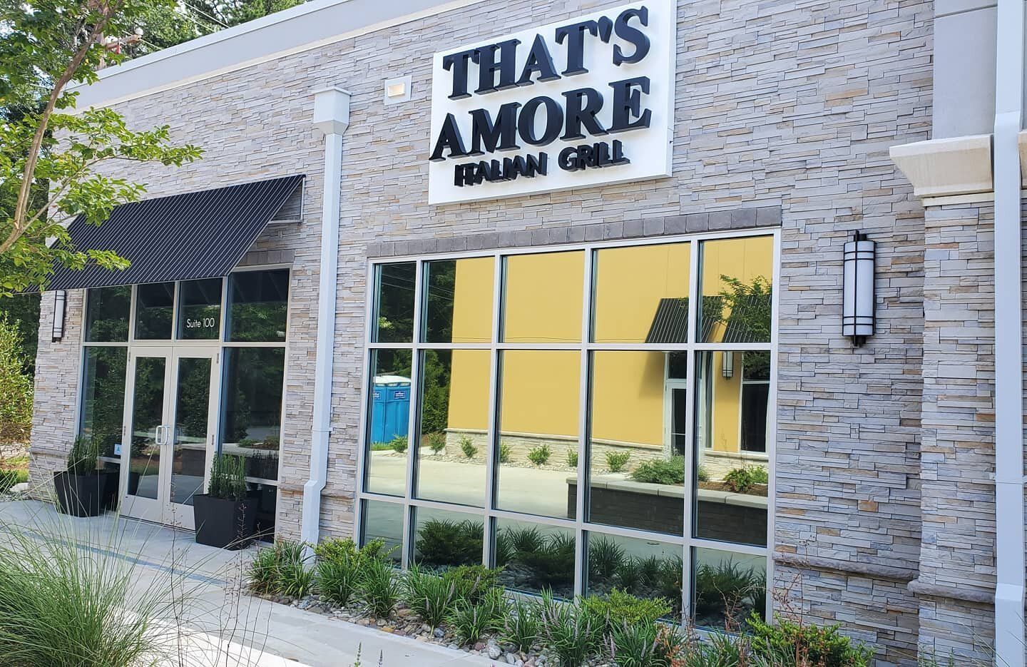 Exterior view of That's Amore Italian Grill, featuring a stone facade, glass windows, and a black striped awning.