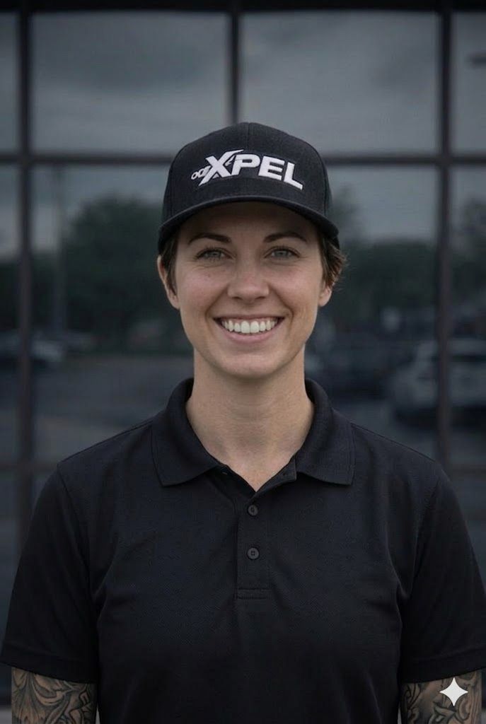 A person wearing a black XPEL baseball cap and a black polo shirt, smiling in front of a dark windowed building.