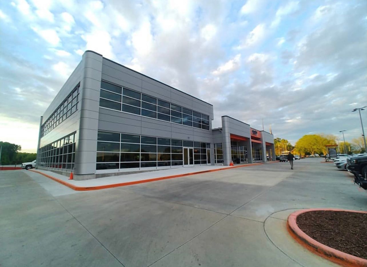 A modern, grey two-story commercial building with large glass windows under a cloudy sky, with a paved parking lot.